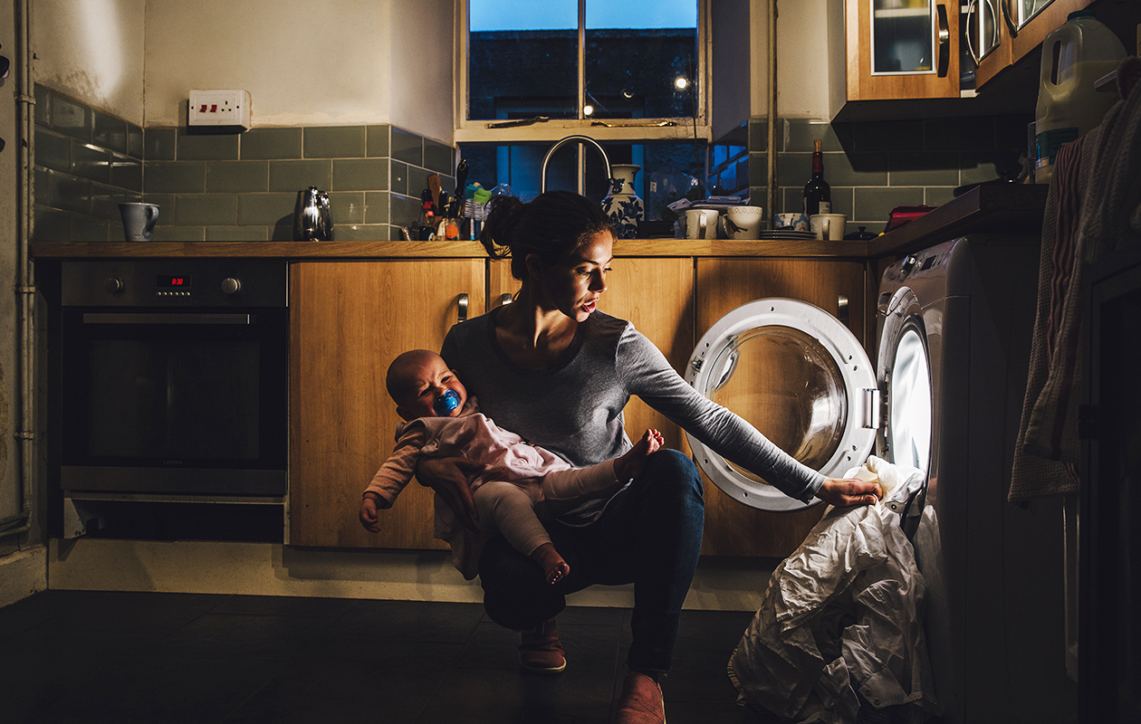 Une jeune femme remplit la machine à laver d'un bras en tenant son bébé de l'autre
