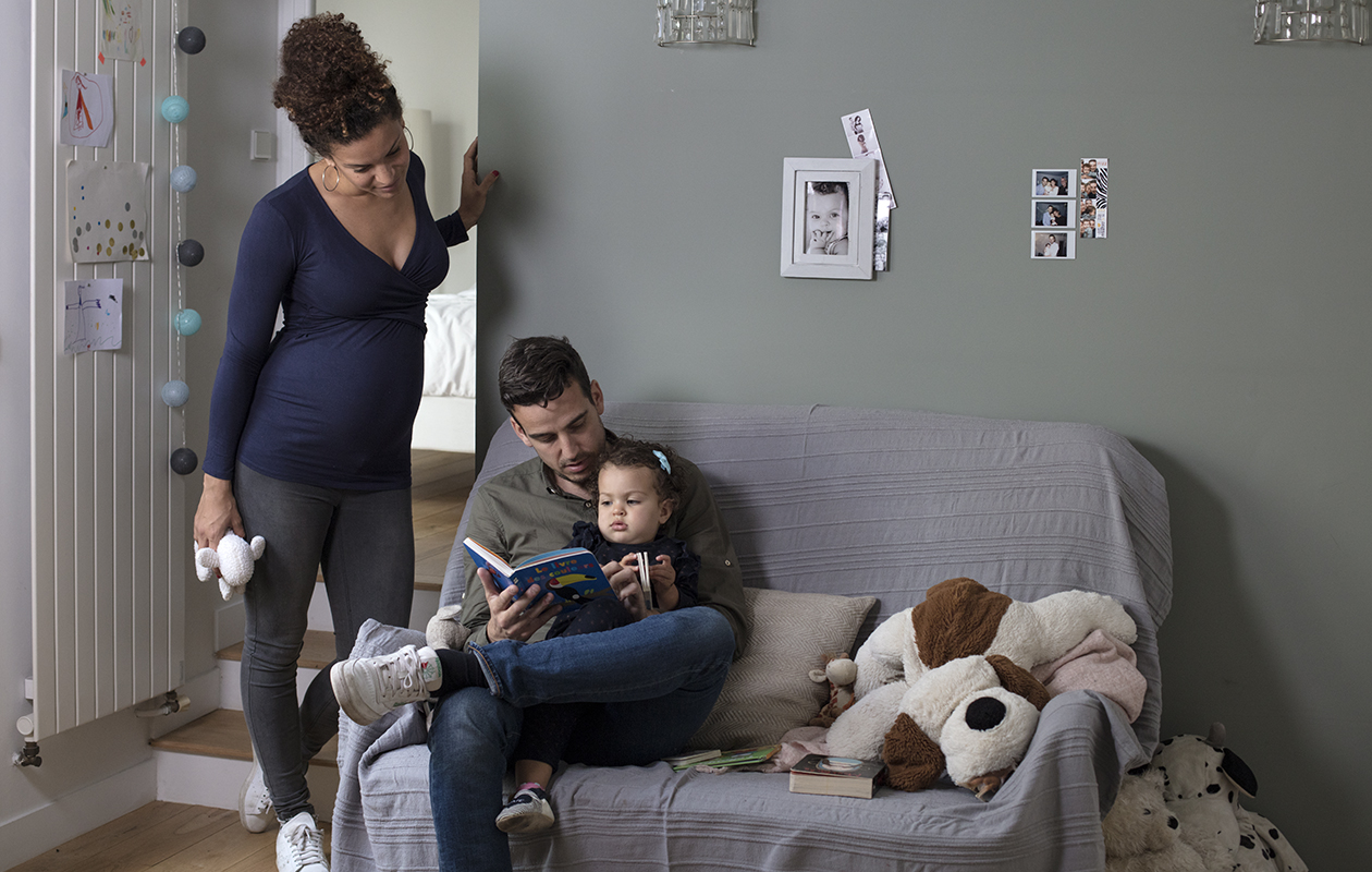 un papa lit une histoire à sa petite fille sous le regard attendri de la maman enceinte