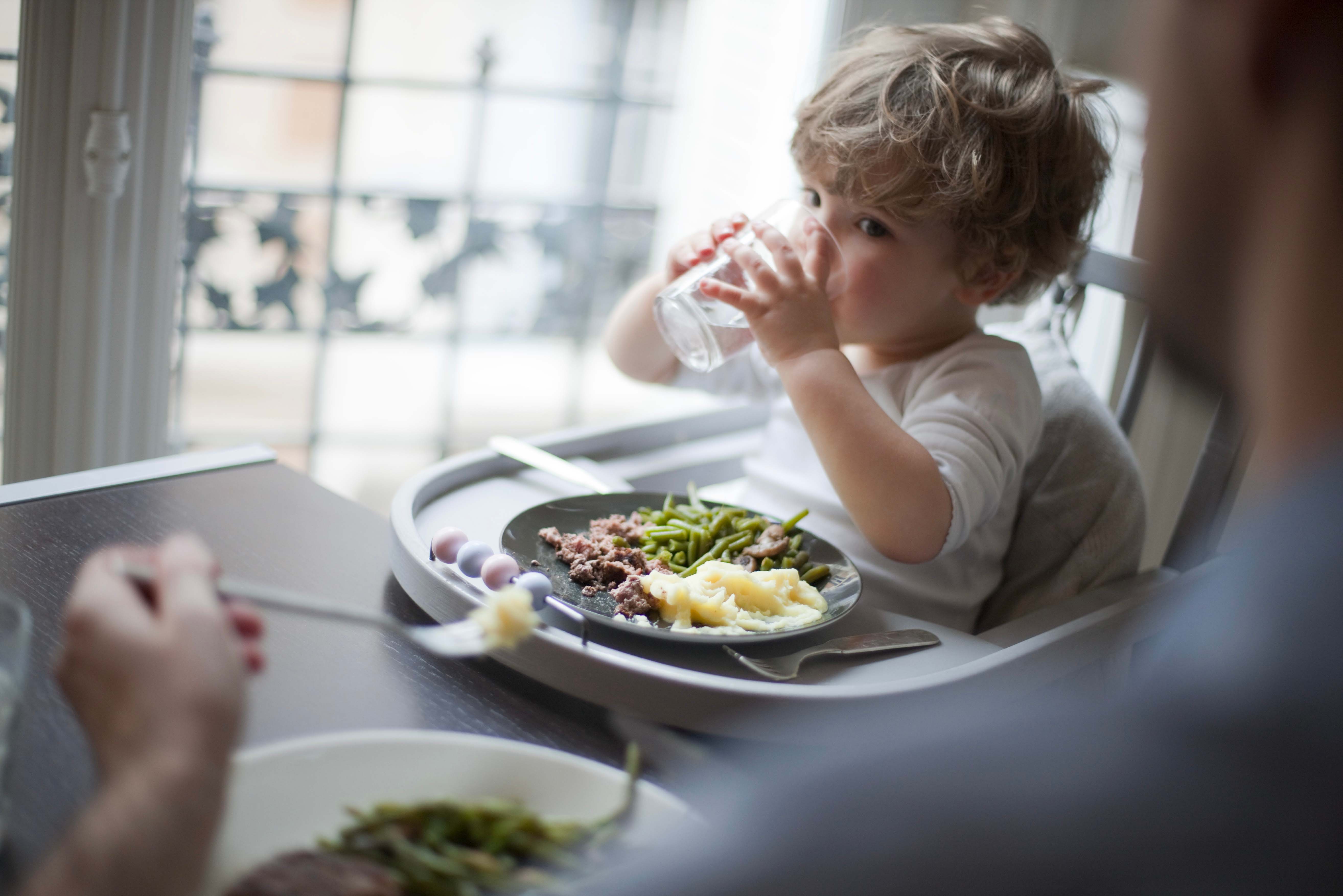 Un jeune enfant boit son verre d'eau devant son assiette, assis dans sa chaise bébé