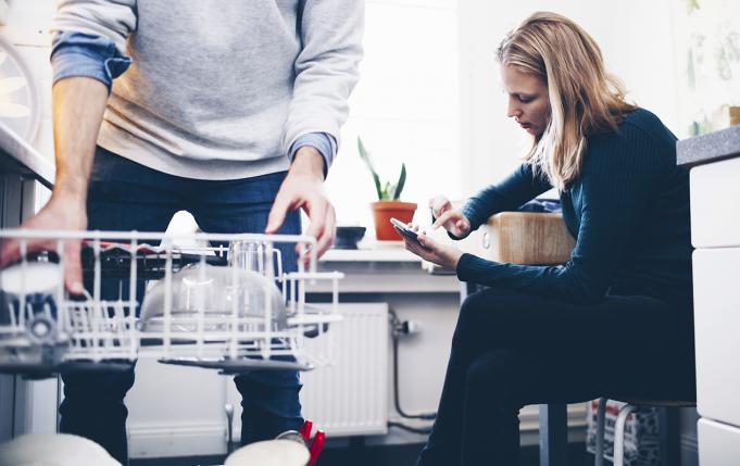 Couple dans la cuisine, la femme est sur son portable pendant que son compagnon remplit le lave vaisselle