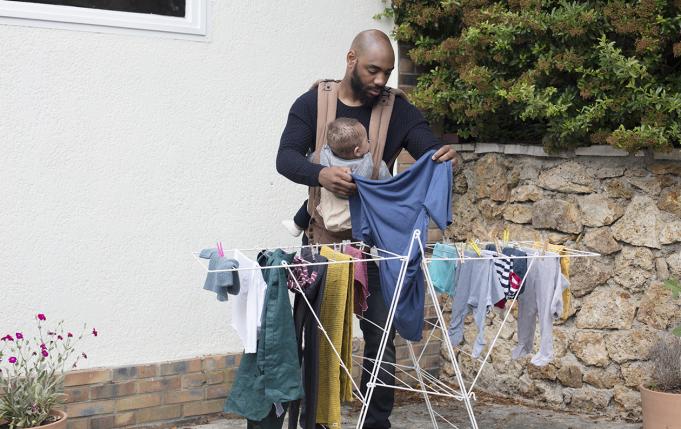 Un jeune papa étend le linge dans son jardin, son bébé contre lui dans un porte-bébé