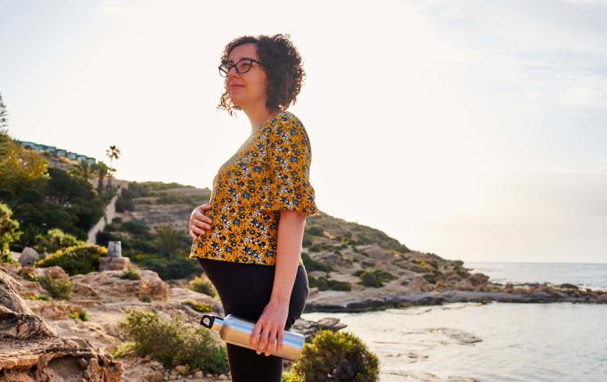 Une femme enceinte porte sa gourde d'eau et se touche le ventre en bord de mer