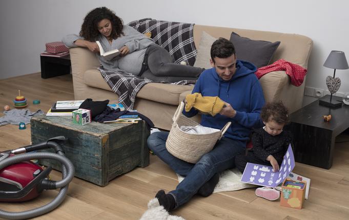 une femme enceinte lit sur le canapé pendant que son compagnon plie du linge à côté de leur petite fille